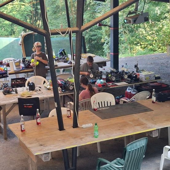 Mehrere Personen arbeiten in einem Außenbereich an verschiedenen RC-Fahrzeugen. Mehrere Tische sind mit Werkzeugen, Flaschen und anderen Utensilien übersät. Bäume und ein Zaun sind im Hintergrund zu sehen.
