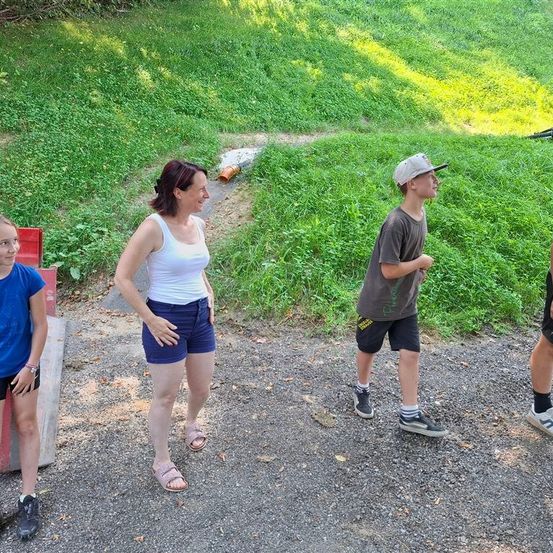 Vier Menschen stehen draußen auf einem Schotterweg. Eine Frau in einem weißen Tanktop und blauen Shorts steht neben einem jungen Jungen, der eine Kappe trägt. Ein Mädchen in einem blauen Shirt steht neben einem roten Objekt. Ein Junge, der Turnschuhe trägt, steht rechts.