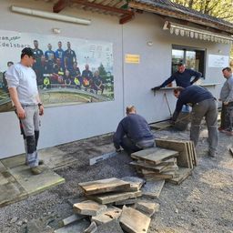 Fünf Männer arbeiten vor einem Gebäude. Einer steht und drei knien, einer benutzt ein Werkzeug. Ein Poster mit Leuten und einem Logo ist an der Wand.