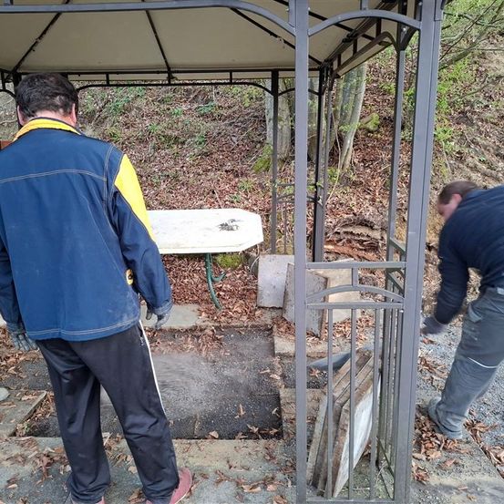 Zwei Männer in Arbeitskleidung stehen neben einem Pavillon, einer beugt sich nach unten, während der andere zusieht. Das Gebiet ist mit Blättern und Steinen bedeckt. Eine Bank steht in der Nähe.