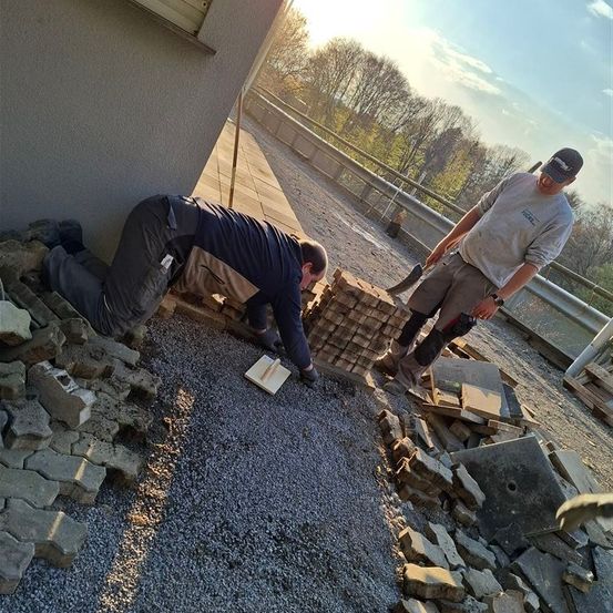 Zwei Männer arbeiten im Freien, einer kniet mit einem Klemmbrett und der andere steht mit einer Kelle. Sie sind von Ziegeln und Schutt umgeben, mit einem Zaun und Grünpflanzen im Hintergrund.