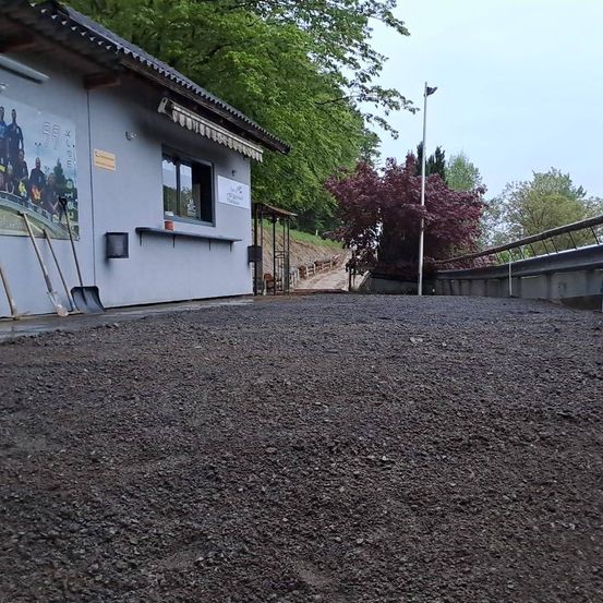 Ein Außenbereich mit Kiesboden, mit einem kleinen Gebäude, einem Poster und einem Vordach. In der Nähe befinden sich Schaufeln und ein Mülleimer. Bäume und ein Pfosten sind im Hintergrund zu sehen.