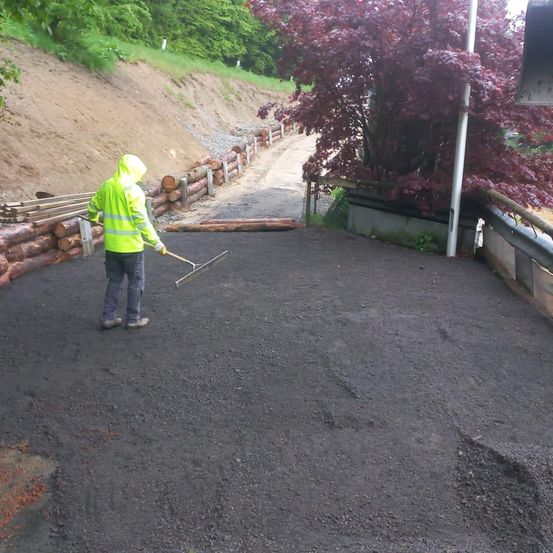 Ein Arbeiter in einer gelben Jacke und gelben Handschuhen glättet die Oberfläche eines Kiesweges mit einem Rechen. Ein buschiger Pflanzen mit violetten Blättern und ein Zaun sind in der Nähe.