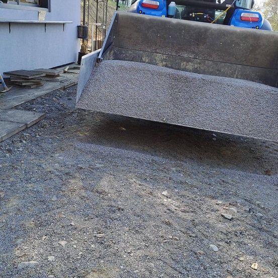 Ein großer blauer Baufahrzeug kippt Kies auf einen Einfahrtsweg. Ein Gebäude ist links, mit Haufen von Kies und einem kleinen Felsen in der Nähe.