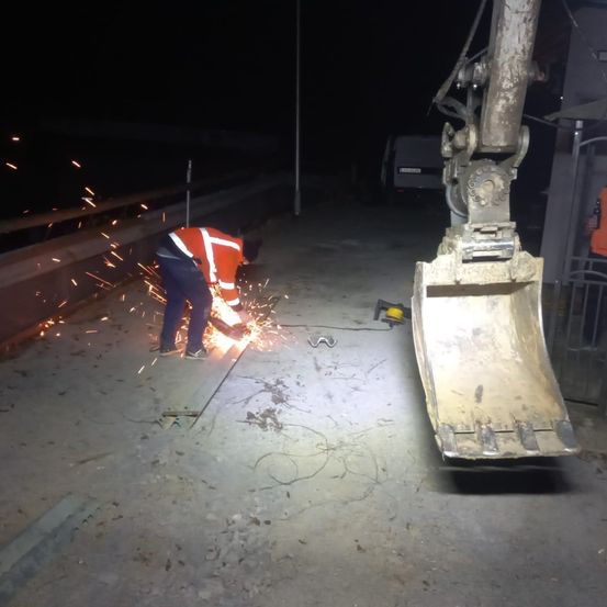 Ein Arbeiter in einer orangefarbenen Sicherheitskleidung schneidet nachts mit einem Schleifer Metall. Funken fliegen. In der Nähe steht ein großes Maschinenteil und ein Lieferwagen.