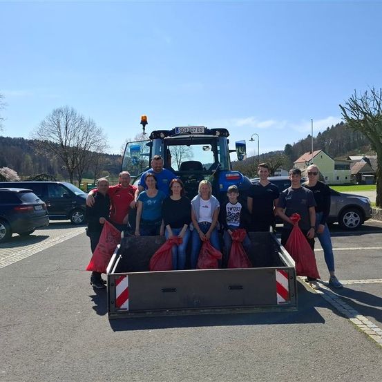 Eine Gruppe von Menschen vor einem blauen Traktor mit roten Säcken, posiert für ein Foto auf einem Parkplatz.