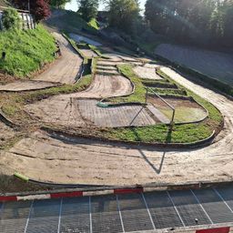 Eine Draufsicht auf eine kurvenreiche Erdbahn, möglicherweise eine Go-Kart-Bahn, mit einer Person, die daran arbeitet. Die Bahn hat Kurven, und der Boden ist mit Erde bedeckt. Ein Metallzaun verläuft entlang der Seite.