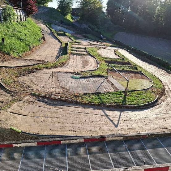 Eine Draufsicht auf eine kurvenreiche Erdbahn, möglicherweise eine Go-Kart-Bahn, mit einer Person, die daran arbeitet. Die Bahn hat Kurven, und der Boden ist mit Erde bedeckt. Ein Metallzaun verläuft entlang der Seite.