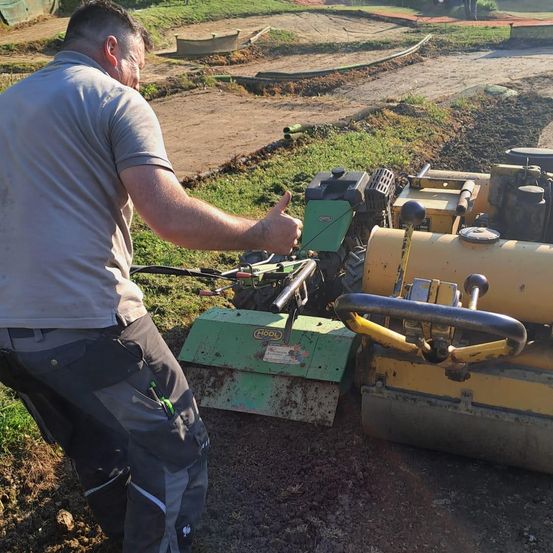 Ein Mann in Arbeitskleidung bedient einen grün-gelben Traktor auf einem schlammigen Feld und zeigt auf die Maschine.