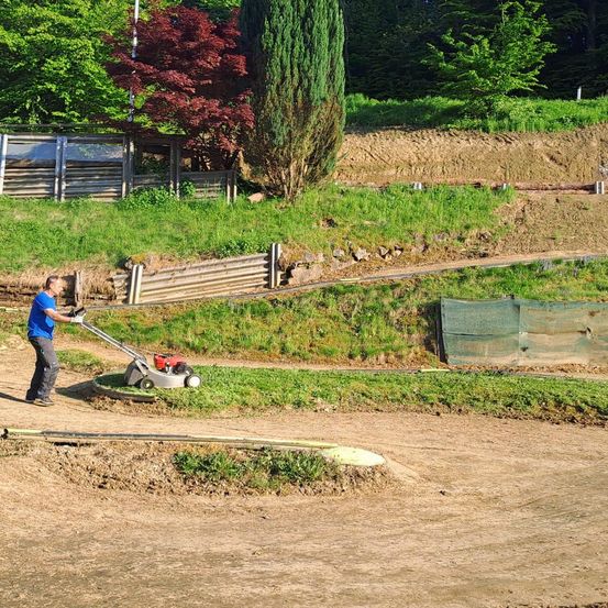 Ein Mann in einem blauen Shirt mäht Gras mit einem roten Rasenmäher auf einem unbefestigten Weg, mit einer Hecke im Hintergrund.