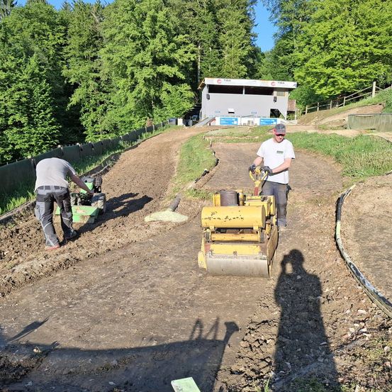 Zwei Männer arbeiten auf einer unbefestigten Straße, einer bedient eine gelbe Maschine und der andere eine grüne, unter einem klaren Himmel.