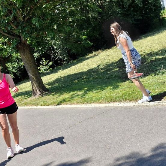 Zwei Frauen stehen auf einem gepflasterten Weg. Eine Frau rennt und hält einen Tennisschläger, während die andere Frau steht und lächelt.