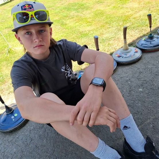 Ein junger Junge sitzt auf einem Betonweg mit drei Curlingsteinen hinter ihm. Er trägt ein schwarzes T-Shirt, schwarze Shorts und Turnschuhe. Er trägt auch eine Kappe mit Logo und Sonnenbrille. Er schaut nach links.