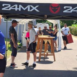 Menschen versammeln sich unter einem Baldachin mit dem 24MX-Logo und führen Gespräche. Ein Tisch mit Getränken steht in der Mitte. In der Nähe hält ein Mann einen roten Lautsprecher.