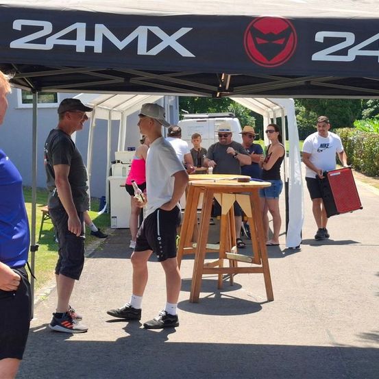 Menschen versammeln sich unter einem Baldachin mit dem 24MX-Logo und führen Gespräche. Ein Tisch mit Getränken steht in der Mitte. In der Nähe hält ein Mann einen roten Lautsprecher.