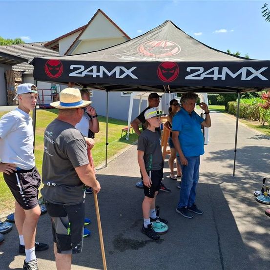 Unter einem Zelt mit 24MX-Marke versammeln sich an einem sonnigen Tag eine Gruppe von Menschen, darunter ein erwachsener Mann mit einem Gehstock. Ein Junge steht vorne, und andere stehen dahinter. Ein Mann schaut nach oben.