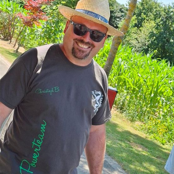 Ein Mann mit Strohhut und Sonnenbrille steht lächelnd im Freien. Er trägt ein graues Shirt mit 'Power Team' in Grün und einem weißen Design. Dahinter ist ein üppiger grüner Hintergrund mit Bäumen und Pflanzen.