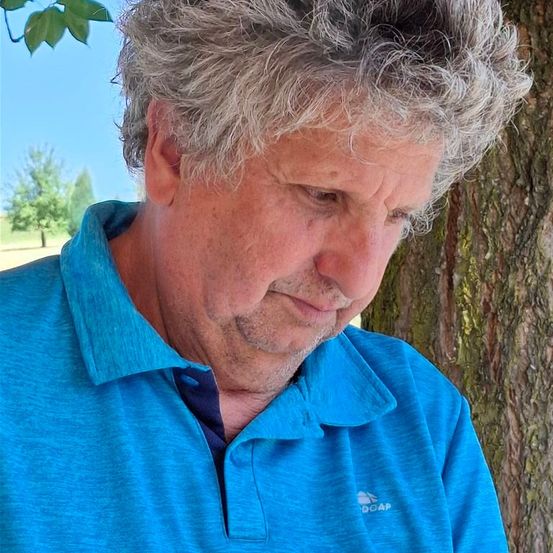 Ein älterer Mann mit grauem Haar trägt ein blaues Poloshirt und schaut nach unten in einer Outdoor-Umgebung. Er steht unter einem Baum, wahrscheinlich in einem Park.