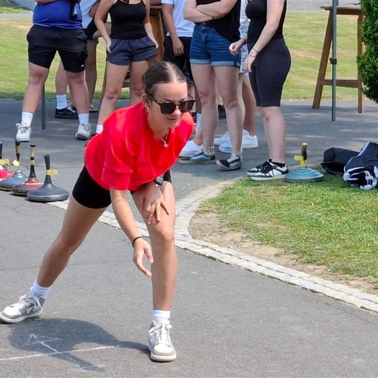 Eine Frau in einem roten Shirt bereitet sich darauf vor, einen Ball zu werfen, während andere zuschauen. Das Spiel beinhaltet das Werfen von Bällen auf Objekte auf dem Boden.