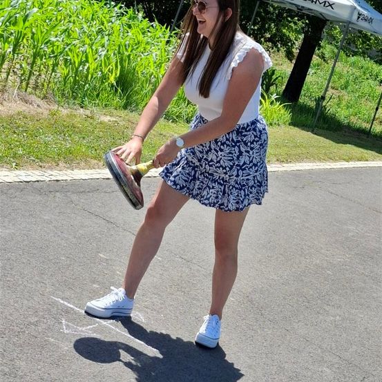 Eine Frau in einer kurzen Hose und weißen Turnschuhen hält einen Schläger, während sie auf einer Straße steht. Hinter ihr befindet sich ein Rasen mit Pflanzen.