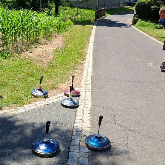 Eine Person steht auf einem Bürgersteig mit vier blauen und silbernen Objekten. Der Gehweg ist von Gras und einem kleinen Pfad gesäumt. Dahinter befindet sich ein Grasfeld mit Mais.