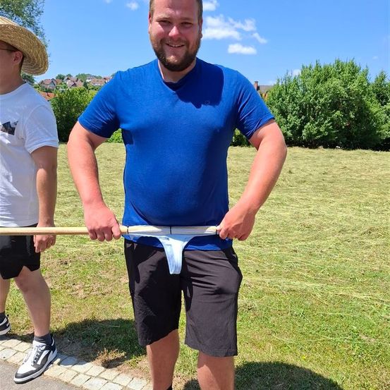 Ein Mann in einem blauen T-Shirt hält einen Stock mit einem weißen Tuch und steht auf einem Grasfeld mit einem anderen Mann, der einen Strohhut trägt.