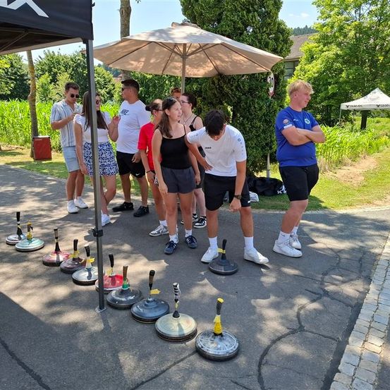 Eine Gruppe von Menschen steht unter einem Regenschirm. Sie stehen vor einer Reihe farbiger Kegel auf dem Boden. Ein Mann beugt sich vor.