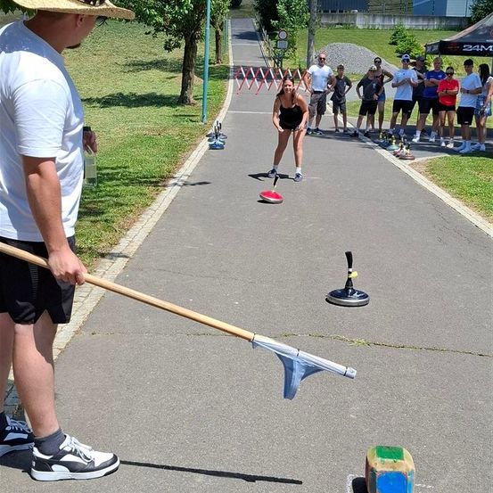 Eine Frau übt auf einem gepflasterten Weg für einen Wurfwettbewerb, während Zuschauer zusehen und ein Mann in der Nähe einen Besen hält.
