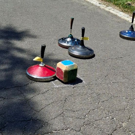 Vier Metallgegenstände mit Griffen liegen auf einer rissigen Asphaltstraße, einer ist rot, zwei sind silbern und einer ist blau. Ein Würfel mit roten und grünen Seiten ist im Vordergrund.