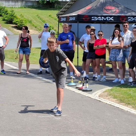 Eine Gruppe von Menschen ist im Freien versammelt, wobei ein Junge in der Mitte steht und möglicherweise an einem Spiel teilnimmt. Zuschauer beobachten, und ein Zelt mit dem Logo 24MX ist in der Nähe.