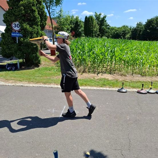 Ein junger Mensch steht auf einer Straße und wirft einen Frisbee. Vier Kegel sind im Vordergrund. Bäume und ein Feld sind im Hintergrund.
