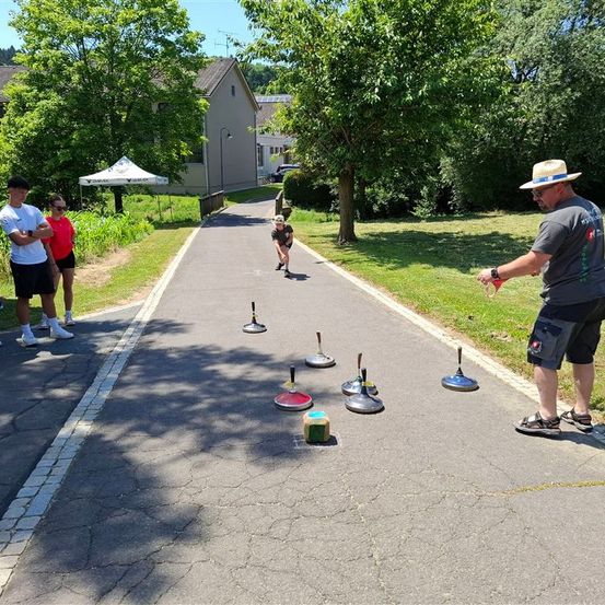 Eine Gruppe von Menschen spielt ein Spiel auf einem gepflasterten Weg mit bunten Objekten und einem Zelt in der Nähe.