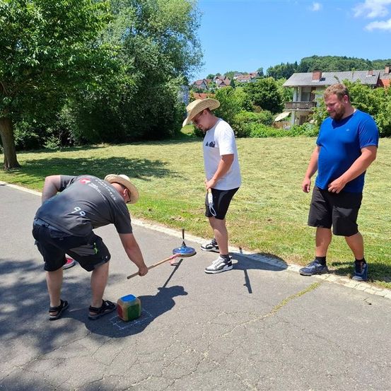 Drei Männer spielen ein Spiel auf der Straße. Einer beugt sich vor, hält einen Hammer und zielt auf einen Würfel. Ein anderer Mann steht hinter ihm, und der dritte steht an der Seite. Bäume und Häuser sind im Hintergrund.