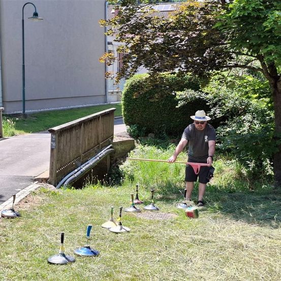 Ein Mann spielt Rasenkegel auf einem Rasen, mit einer Brücke und einem Gebäude im Hintergrund.