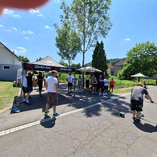 Unter einem Baldachin bereitet sich eine Gruppe von Menschen auf eine Veranstaltung vor, einige stehen herum und ein Mann übt auf der Straße. Bäume und Häuser sind im Hintergrund.