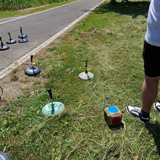 Eine Person steht auf Gras neben einem Weg mit mehreren Metallobjekten, darunter ein großer Ring und ein Würfel, die Schatten auf den Boden werfen.