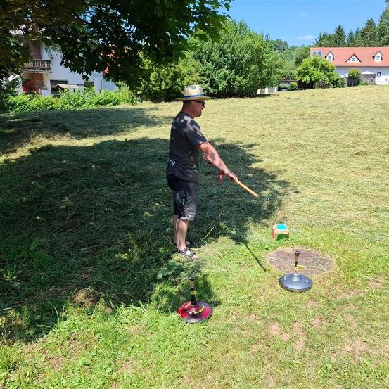 Ein Mann steht auf einem Rasenfeld und hält einen Stock in der Hand, um einen Wurf zu machen. Zwei Zielscheiben liegen auf dem Boden. Ein Haus und Bäume sind im Hintergrund.