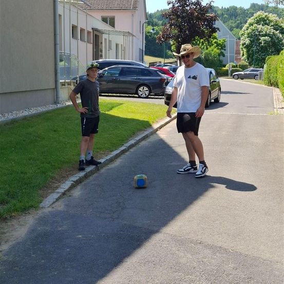 Zwei Männer stehen auf einer Straße, einer trägt einen Cowboyhut und der andere eine Kappe. Ein Fußball liegt auf der Straße. Hinter ihnen sind auf beiden Seiten der Straße Autos geparkt.