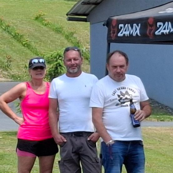 Drei Personen stehen zusammen vor einem Schuppen mit einem Vordach. Die Frau trägt ein rosa Tanktop und schwarze Shorts. Die Männer tragen weiße Hemden und einer hält eine Flasche.