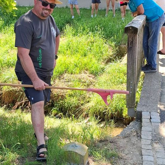 Ein Mann in Hut und Sonnenbrille fängt mit einem Fischernetz in einem kleinen Bach. Hinter ihm lehnt sich ein Mann über einen Holzpfosten. Mehrere Personen in Shorts stehen im Hintergrund in der Nähe eines Gebäudes.