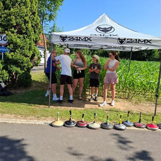 Eine Gruppe von Menschen steht unter einem weißen Zelt mit dem 24MX-Logo neben einem Maisfeld. Sie tragen Turnschuhe und einige halten Flaschen.