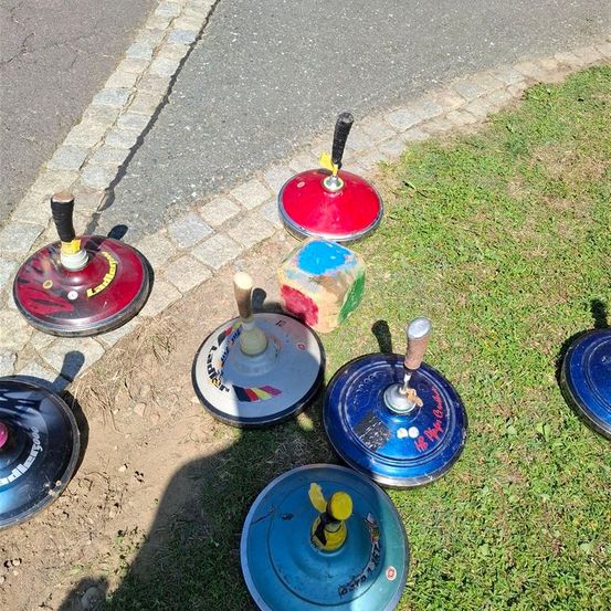 Mehrere Spinnteller sind auf Gras neben einem gepflasterten Weg platziert. Sie sind in verschiedenen Farben bemalt und haben Griffe. Ein Schatten wird auf das Gras geworfen.