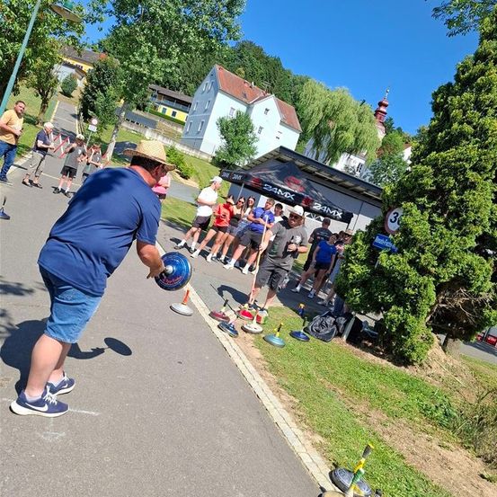 Ein Mann wirft einen Ball auf ein Croquet-Set auf der Straße. Zuschauer beobachten. In der Nähe ein Zelt mit dem Schild 24MX. Bäume und ein Haus im Hintergrund.