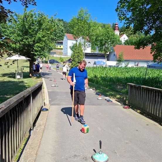 Ein Mann übt das Werfen eines Balls auf ein Ziel an einem sonnigen Tag, mit mehreren Häusern und vorbeigehenden Personen im Hintergrund.