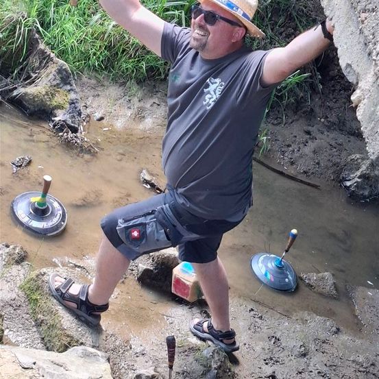 Ein Mann steht in schlammigem Wasser, trägt einen Hut und eine Sonnenbrille, mit drei Werkzeugen in der Nähe.