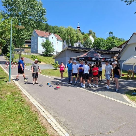 Eine Gruppe von Menschen übt eine Sportart auf der Straße, mit einem Zelt und einigen Häusern in der Nähe. Sie stehen vor einem Zelt mit einem Banner, auf dem 24MX steht.