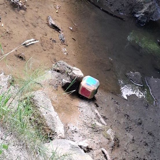 Ein schmutziger Flussufer mit einem kleinen, farbenfrohen, würfelförmigen Objekt in der Nähe des Wasserrandes. Die Umgebung ist schlammig mit verstreuten Abfällen.