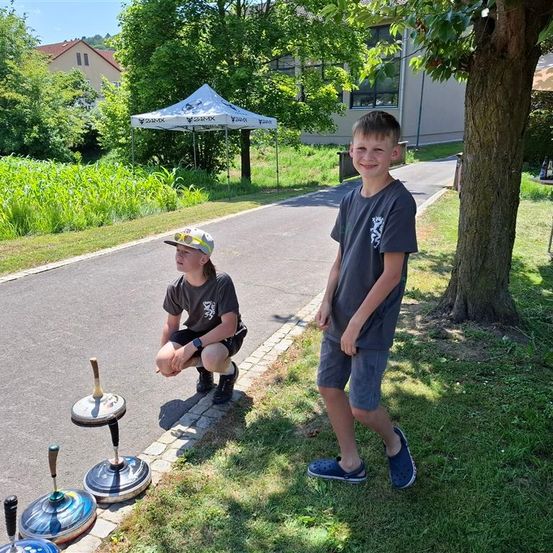 Zwei Jungen stehen draußen mit drei großen Drehscheiben. Ein Junge hockt und der andere steht lächelnd. Dahinter ist ein Rasen mit einem Baum und einem weißen Zelt.