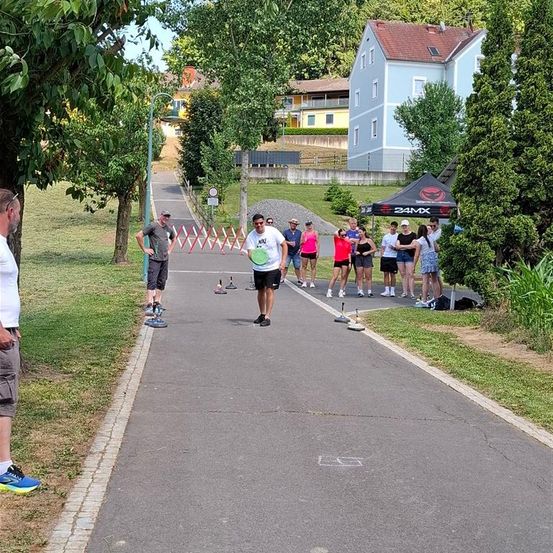 Eine Gruppe von Menschen steht auf einem Weg in einem Park. Einige tragen Turnschuhe und andere sind barfuß. Sie schauen einen Mann an, der einen Frisbee hält. In der Ferne befindet sich ein Zelt und ein Haus.
