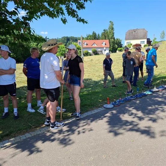 Eine Gruppe von Menschen versammelt sich auf einem Rasen. Einige tragen Hüte und Turnschuhe, und ein Mann hält einen Stock. Im Hintergrund befinden sich Häuser, Bäume und ein klarer Himmel.